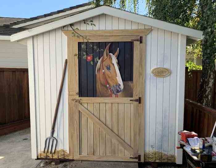 A building with a hand-painted mural on the front depicting a horse looking out of a stable, surrounded by realistic details like a pitchfork and an apple.