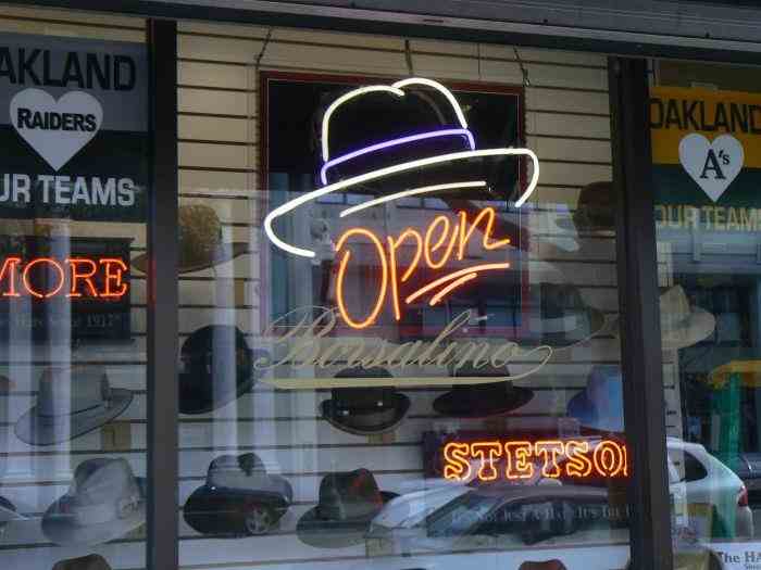 Storefront window display featuring a neon 'Open' sign shaped like a hat, with the brand name 'Borsalino' below it, surrounded by various hats and other neon signs.