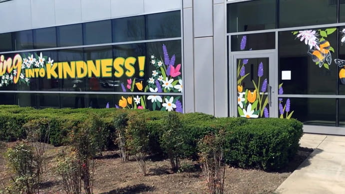 Hand-painted window mural spanning the glass front of a Kaiser hospital building, with spring flowers and the word Kindness