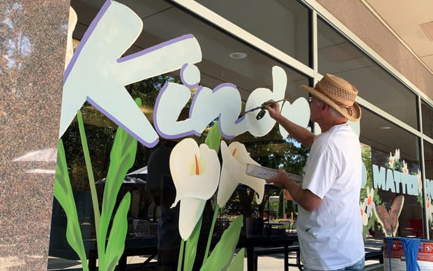 Tim Taylor in a straw hat painting the word Kindness by hand on a large window, brush in one hand and paint tray in the other, with hand-painted calla lilies below the lettering